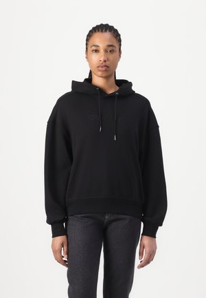 Black hooded sweatshirt with a loose fit, featuring a soft texture and embroidered logo in the center. Paired with dark jeans.