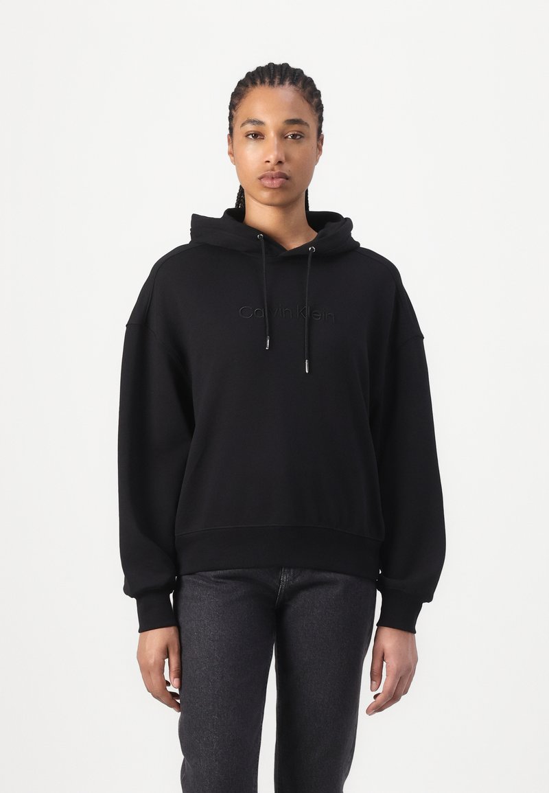 Zwarte hooded sweatshirt met een losse pasvorm, voorzien van een zachte textuur en een geborduurd logo in het midden. Gepaarde met donkere jeans.