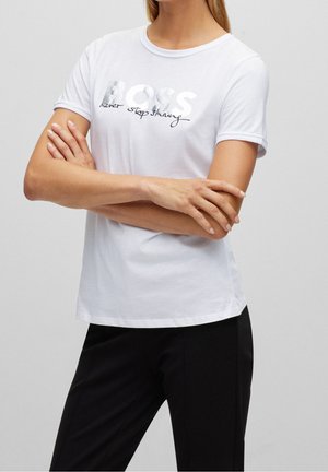 Kobieta w białej koszulce z srebrnym napisem „BOSS” oraz czarnym napisem „never stop shining”, ze skrzyżowanymi ramionami, ubrana w czarne spodnie.