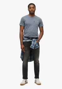 Camiseta de algodón azul con un bolsillo en el pecho, combinada con jeans oscuros y una camisa de cuadros atada a la cintura. Zapatillas blancas con detalles en gris.