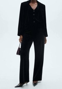 Costume en velours noir avec un blazer cintré, un pantalon à jambes larges et un haut boutonné. Escarpins brillants à bout pointu et petit sac à main en motif crocodile.