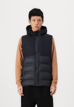 Black puffer vest with hood, featuring a snap and zipper closure, quilted design, and a zippered chest pocket, worn over a brown long-sleeve top.