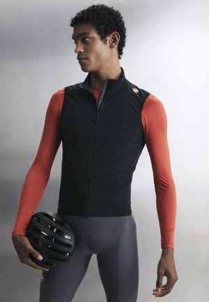 Uomo con gilet da ciclismo nero senza maniche, maglia rossa a maniche lunghe, pantaloncini da ciclismo grigi, che tiene un casco nero, in piedi davanti a uno sfondo semplice.