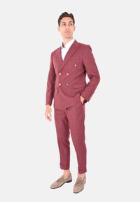 Abito bordeaux doppiopetto con bottoni dorati, taglio slim, dotato di due tasche frontali e polsini arrotolati, abbinato a mocassini marrone chiaro.