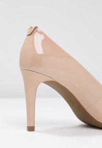 Chaussure à talon haut en cuir verni couleur nude avec un bout pointu, un design élégant, et un petit accent doré à l'arrière près du talon.