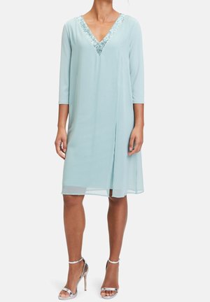 Žena v světle modrých šatech po kolena se tříčtvrtečními rukávy a zdobeným výstřihem do V s flitry, doplněná stříbrnými sandály na vysokém podpatku.