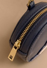 Kulatá modrá taška s texturovaným povrchem, zlatým zipem a kovovými detaily. Detailní pohled zdůrazňuje zip a švy.