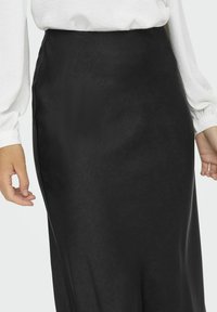 Jupe longue en satin noir avec une texture lisse et une taille ajustée, portée avec un chemisier blanc. Présente une couture latérale pour un design épuré.