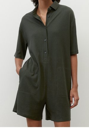 Femme portant une combinaison courte côtelée vert foncé à manches courtes boutonnée avec col et poches, debout avec une main dans la poche.