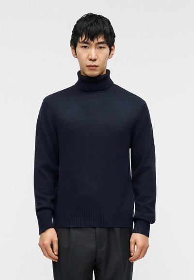 Pull à col roulé bleu marine en maille douce avec poignets et ourlet côtelés, associé à un pantalon gris à fines rayures, posé devant un fond neutre.