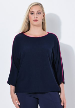 Frau mit langen blonden Haaren, die einen marineblauen Pullover mit pinkem Saum und passende marineblaue Hose trägt, steht vor einem weißen Hintergrund.
