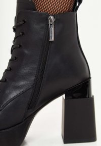 Botín de cuero negro con un prominente tacón cuadrado, cordones en la parte frontal y detalle de cremallera lateral. Se puede ver una malla negra texturizada en la parte superior.