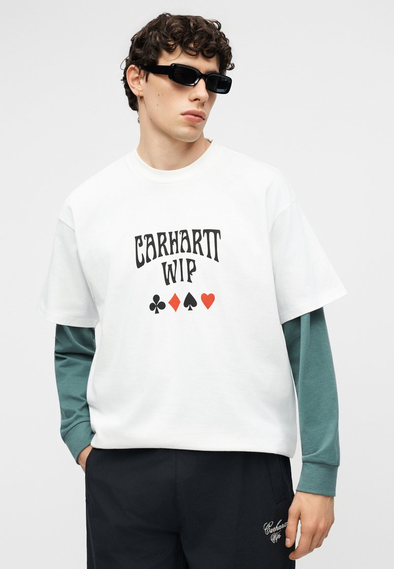 Junger Mann mit schwarzer Sonnenbrille, weißem Carhartt WIP T-Shirt über einem grünen Langarmshirt und schwarzen Hosen mit Logo am Bein.