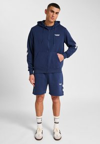 Hoodie zippé marine avec poche kangourou et logo, associé à un short assorti. Les deux présentent des accents en chevrons blancs. Tissu doux et décontracté.