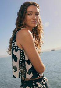 Zwart-wit gestreept crop top met abstracte vormen, details van zijkoordjes en een gestructureerde stof, gedragen door een model dichtbij water.