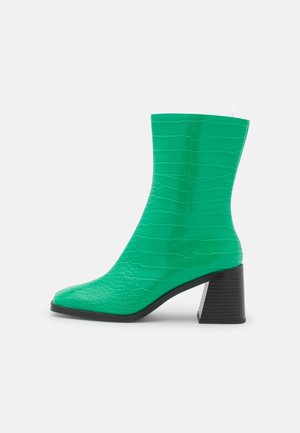 Botte verte mi-mollet à texture crocodile, bout carré et talon bloc noir épais, vue de profil sur fond blanc.