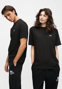 Άνδρας και γυναίκα που φοράνε μαύρα T-shirts της Lacoste και μαύρο παντελόνι, στέκονται δίπλα-δίπλα μπροστά σε ένα απλό ανοιχτό φόντο.