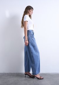 Γυναίκα με μακριά μαλλιά που φοράει λευκό μπλουζάκι με κοντά μανίκια, φαρδιά μπλε τζιν με ψηλή μέση, μαύρα σανδάλια με τακούνι και ασημένια βραχιόλια, στέκεται πλαγίως.