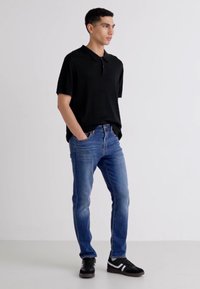 Crna polo majica, plave traperice i crne tene s bijelim detaljima. Model stoji protiv jednolične pozadine, prikazujući ležernu odjeću.