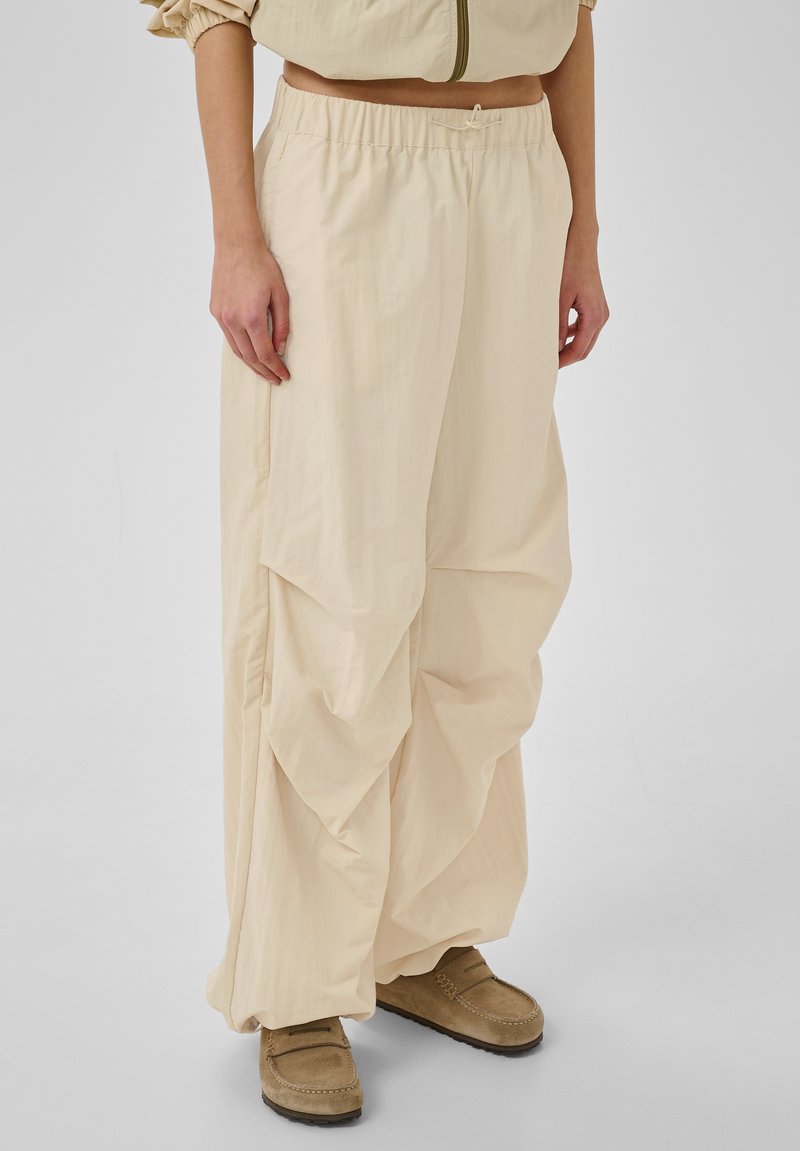 Person trägt lockere beige Hose mit elastischem Bund und braune Mokassins vor einfachem hellem Hintergrund.