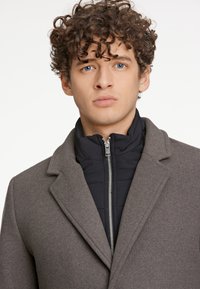Cappotto grigio in lana con rever a vista, indossato sopra una giacca trapuntata blu navy. Presenta una chiusura frontale con zip e una vestibilità strutturata.