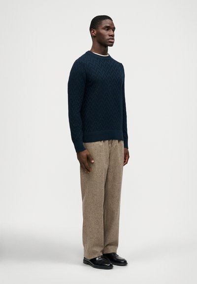 Dunkelblauer Strickpullover mit strukturiertem Muster, kombiniert mit beigen Stoffhosen. Die Passform ist lässig und verfügt über einen gerippten Hals- und Ärmelbund.