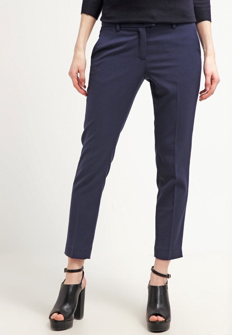 Femme portant un pantalon slim bleu marine coupé à la cheville et des sandales noires à talon bloc et orteils ouverts, debout devant un fond clair uni.