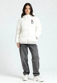 Giacca a vento bianca con zip frontale, cappuccio ed emblema con mano e cuore. Abbinata a jeans grigi e sneaker bianche.