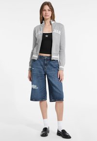Sivý zipsový kardigán s bielym textom "GUESS JEANS", spárovaný s modrými džínovými bermudami s poškodeným vzorom. Čierne mokasíny a biele ponožky.