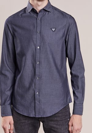 Man draagt een donkerblauw overhemd met lange mouwen, knopen en een klein patroon, gecombineerd met een zwarte spijkerbroek, staand voor een effen achtergrond.