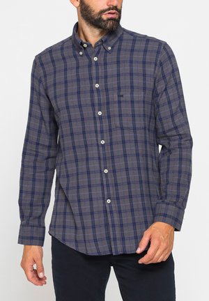 Uomo che indossa una camicia a quadri grigia e blu con bottoni, maniche lunghe e taschino sul petto, abbinata a pantaloni scuri.