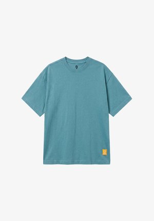 Niebieski t-shirt z krótkim rękawem i okrągłym dekoltem z małą żółtą kwadratową naszywką blisko dolnego brzegu na białym tle.