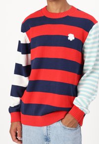 Pull en tricot avec des rayures rouges, bleu marine et blanches, présentant une manche à rayures bleu clair et un petit logo fleur blanche sur la poitrine.