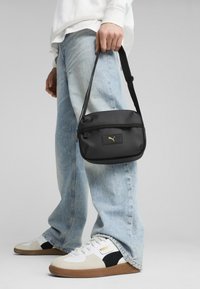 Svart, slätt tyg crossbody-väska med en guldig logotyp, rektangulär form, dragkedjestängning och justerbar rem. Bärs med ljusblå jeans.