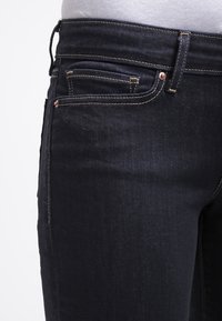 Vaqueros de denim azul oscuro con un corte ajustado. Incluyen herrajes de color cobre, diseño de cinco bolsillos y detalles de costura sutiles.