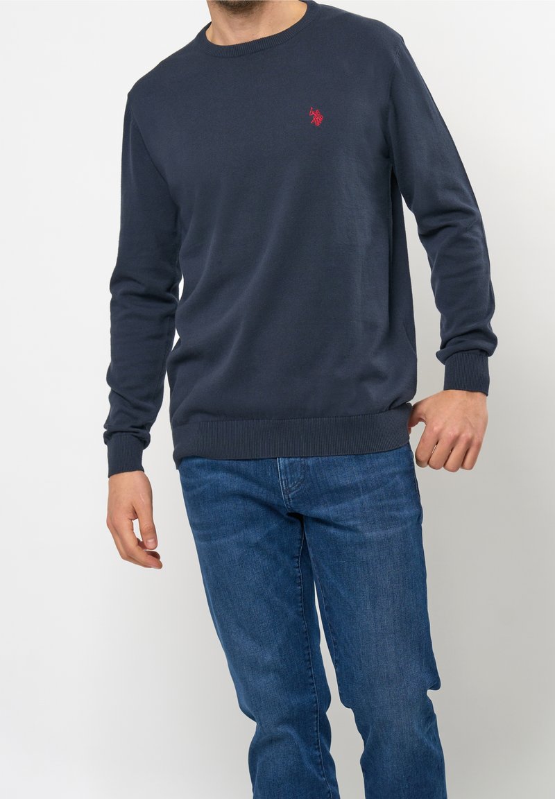 Homme portant un pull bleu marine à manches longues avec un logo rouge sur la poitrine et un jean bleu sur un fond clair uni.