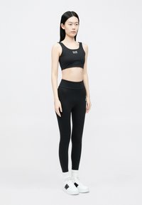 Fekete sportmelltartó logóval, magasderekú fekete leggings, és fehér sportcipő fekete részletekkel. A modell egy sima háttér előtt áll függőlegesen.