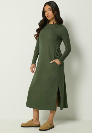 Femme aux longs cheveux foncés portant une robe verte olive à manches longues avec une fente sur le côté et des chaussures beige à enfiler, debout devant un fond uni.