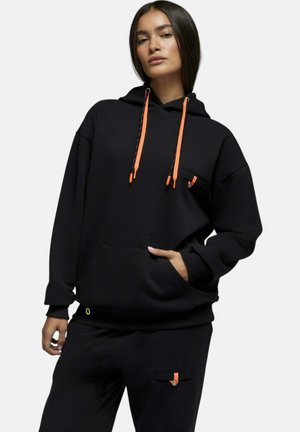 Jeune femme portant un sweat à capuche noir avec des cordons orange et un pantalon noir assorti avec une poche accentuée orange, debout avec une main dans la poche.
