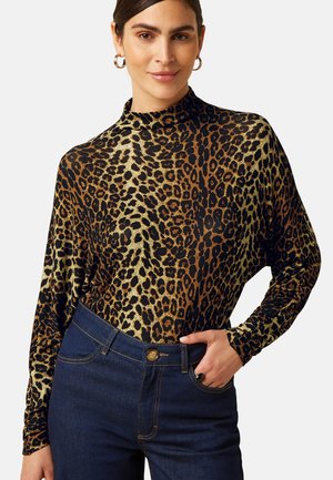 Vrouw draagt een lange mouwen top met luipaardprint en donkerblauwe high-waist jeans met haar rechterhand in de zak.
