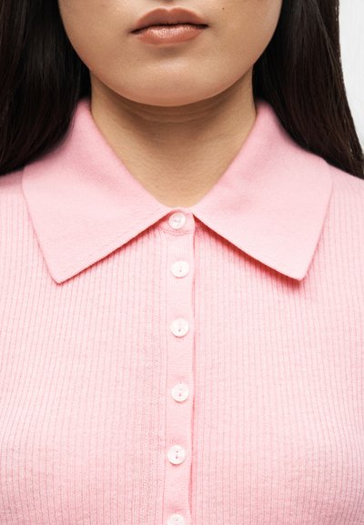 Κοντινή φωτογραφία του κάτω μέρους του προσώπου και του λαιμού ενός ατόμου που φοράει ένα ροζ ριγωτό πουλόβερ με κουμπιά και μεγάλο γιακά.