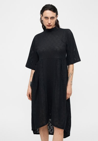 Γυναίκα με σκούρα μαλλιά που φοράει μαύρο, υφασμάτινο φόρεμα μέχρι το γόνατο με κοντά, φαρδιά μανίκια και ψηλό λαιμό, στέκεται μπροστά σε απλό φόντο.