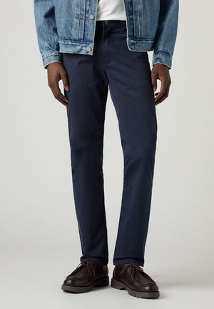 Homme portant un pantalon droit bleu foncé, une chemise blanche, une veste en jean et des chaussures à lacets marron foncé se tenant sur un fond gris.