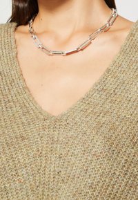 Collana statement in argento con link rettangolari allungati e una sottile texture, indossata su un maglione verde oliva con scollatura a V e texture.