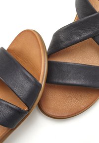 Schwarze Ledersandalen mit breiten Kreuzriemen und einer Innensohle aus tanfarbener Wildleder. Sie haben eine flache Sohle und eine glatte, strukturierte Oberfläche.