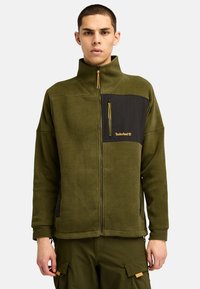 Giacca in pile verde oliva con una tasca sul petto con zip nera, colletto alto e zip gialla. Presenta una texture morbida e una vestibilità rilassata.