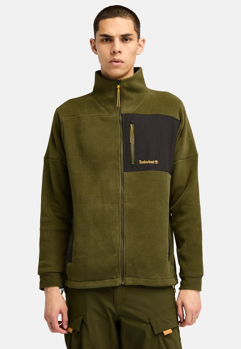 Giacca in pile verde oliva con una tasca sul petto con zip nera, colletto alto e zip gialla. Presenta una texture morbida e una vestibilità rilassata.