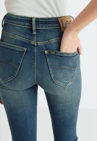 Ciemnoniebieskie dżinsy z wysokim stanem, dwoma tylnymi kieszeniami oraz brązową skórzaną łatą z napisem "Lee" na pasku.