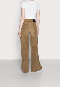 Nainen, jolla on päällään korkeavyötäröiset leveälahkeiset ruskeat manseryhousut, valkoinen lyhyt t-paita ja valkoiset lenkkarit, seisoo valkoista seinää vasten.