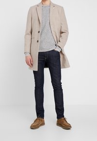 Hellbeige Mantel mit Revers, dunkelgrauer Strickpullover, dunkle Jeans und hellbraune Wildlederschuhe. Saubere Linien und insgesamt schlichtes Design.